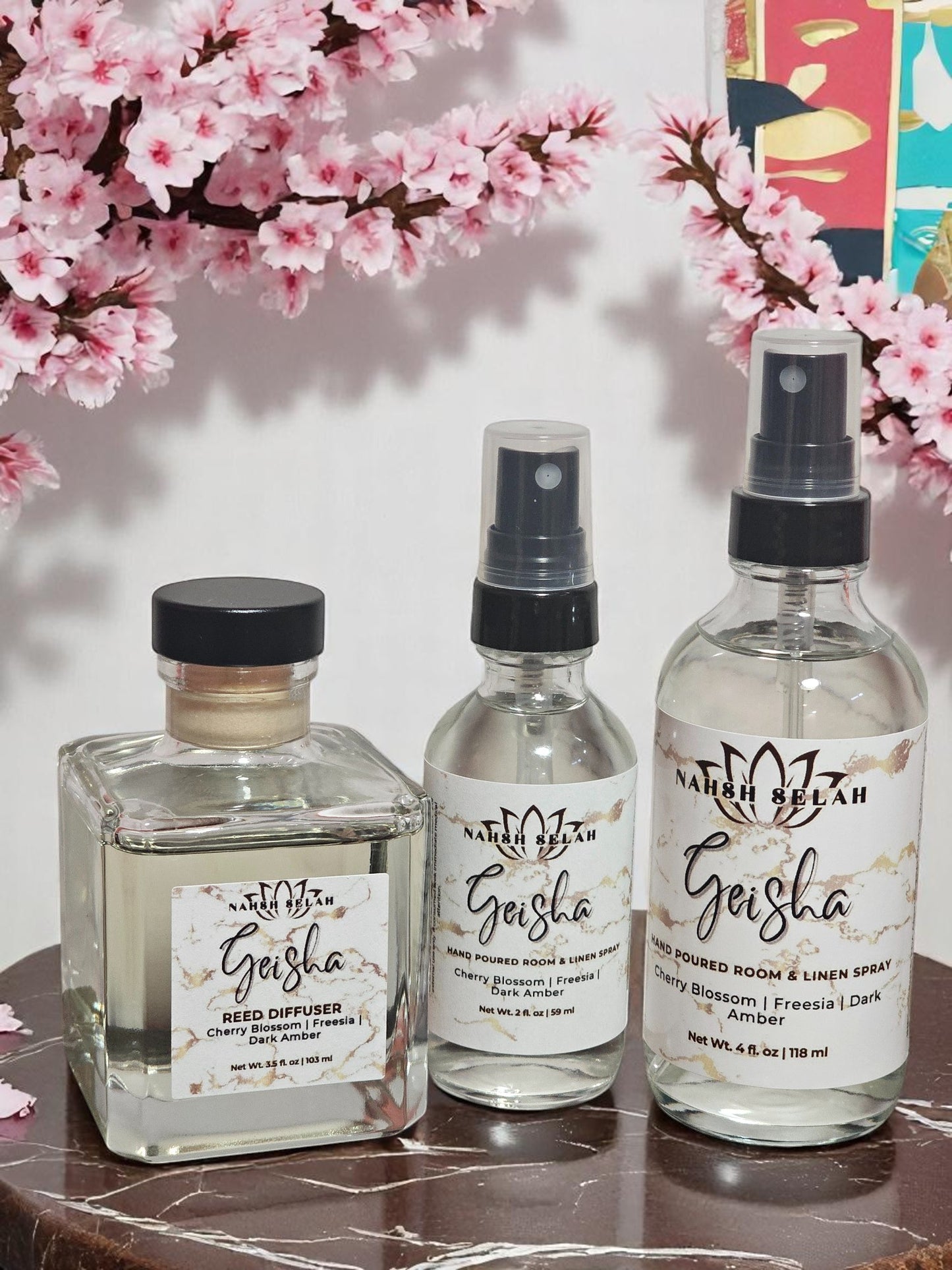 Three Geisha products including a diffuser and two spray bottles on a marble surface with cherry blossoms in the background.