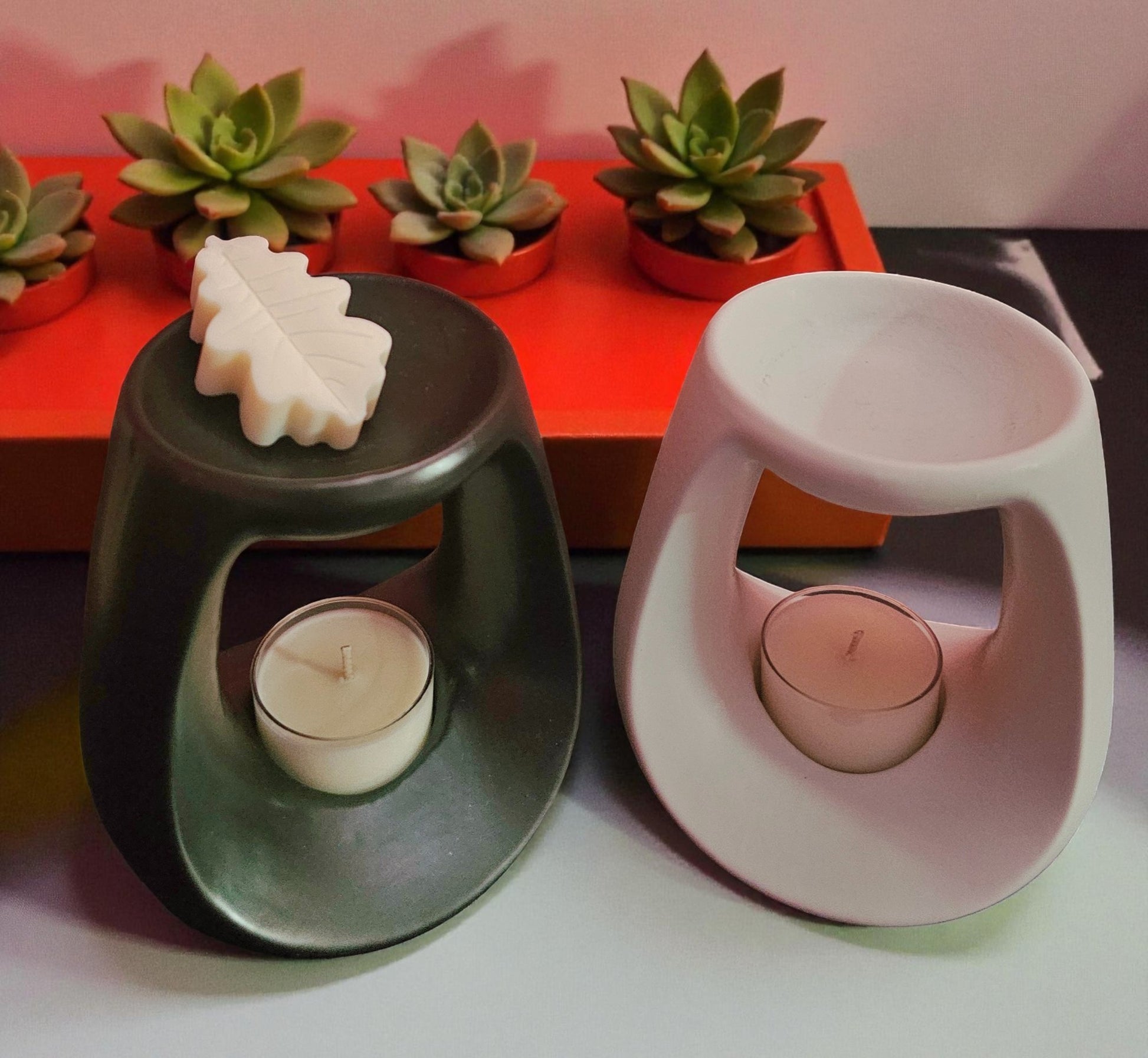 Two candle holders with candles inside, one black and one white, on a surface with succulents in the background.